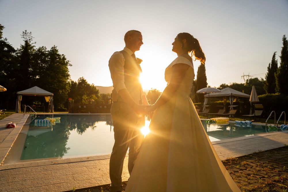 Bride and groom holding hands in golden sunset light by a pool, silhouetted against warm glowing backlight.