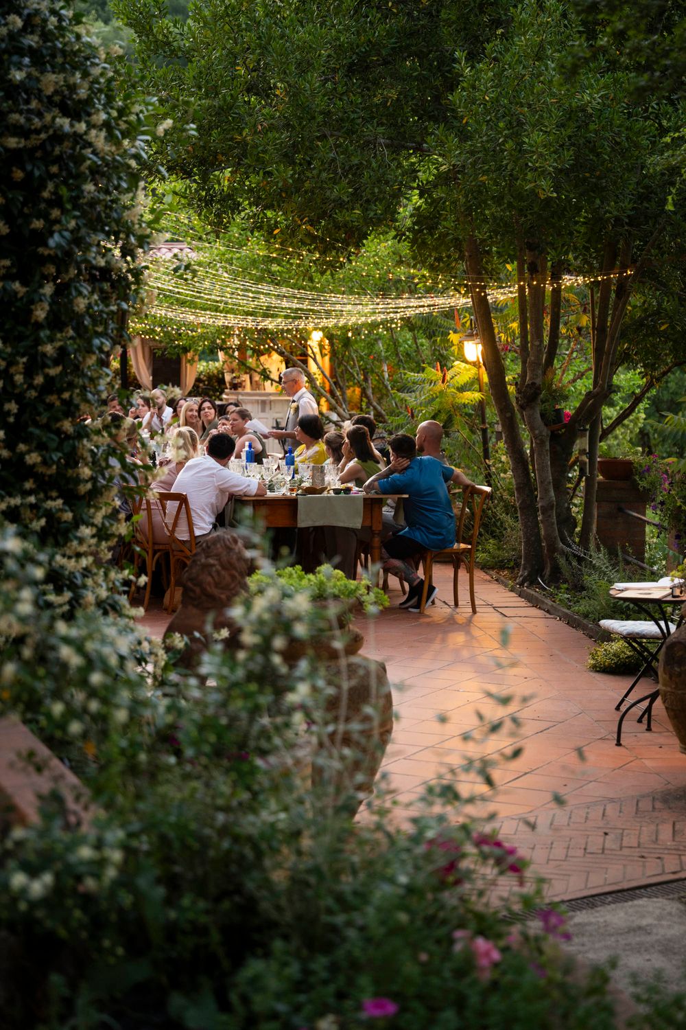 Wedding guests dine at long tables in a lush garden patio adorned with warm string lights at dusk.