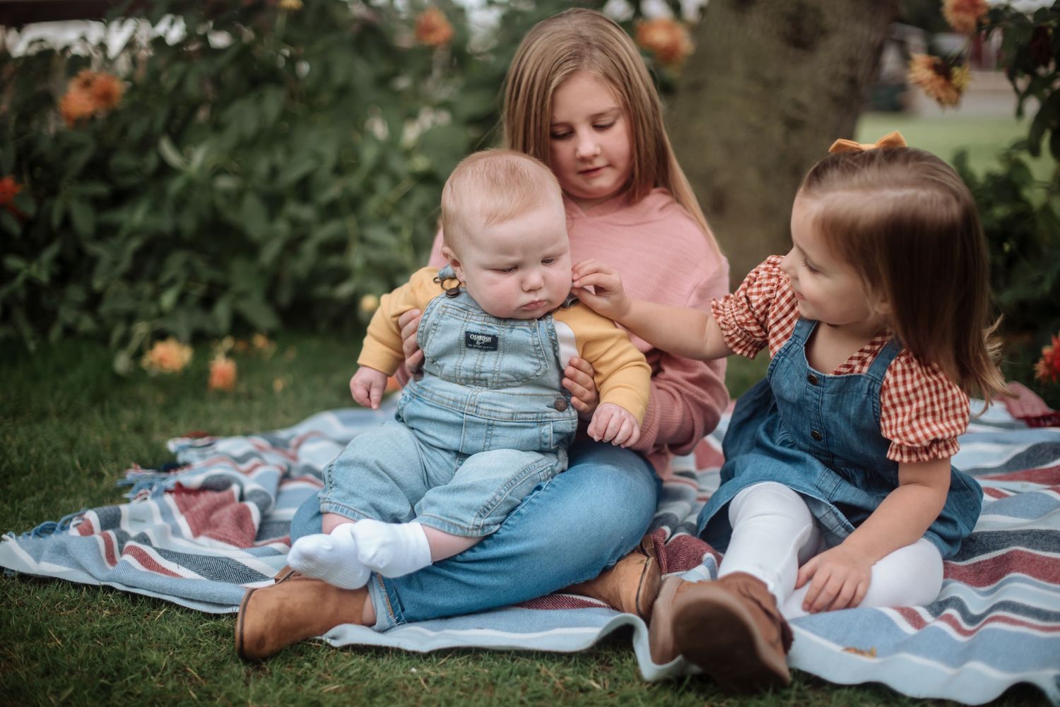 Three children sitting together on a picnic blanket in a garden with blooming flowers.