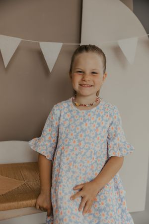 Child wearing a light blue floral print dress poses against a neutral wall with white bunting decorations.