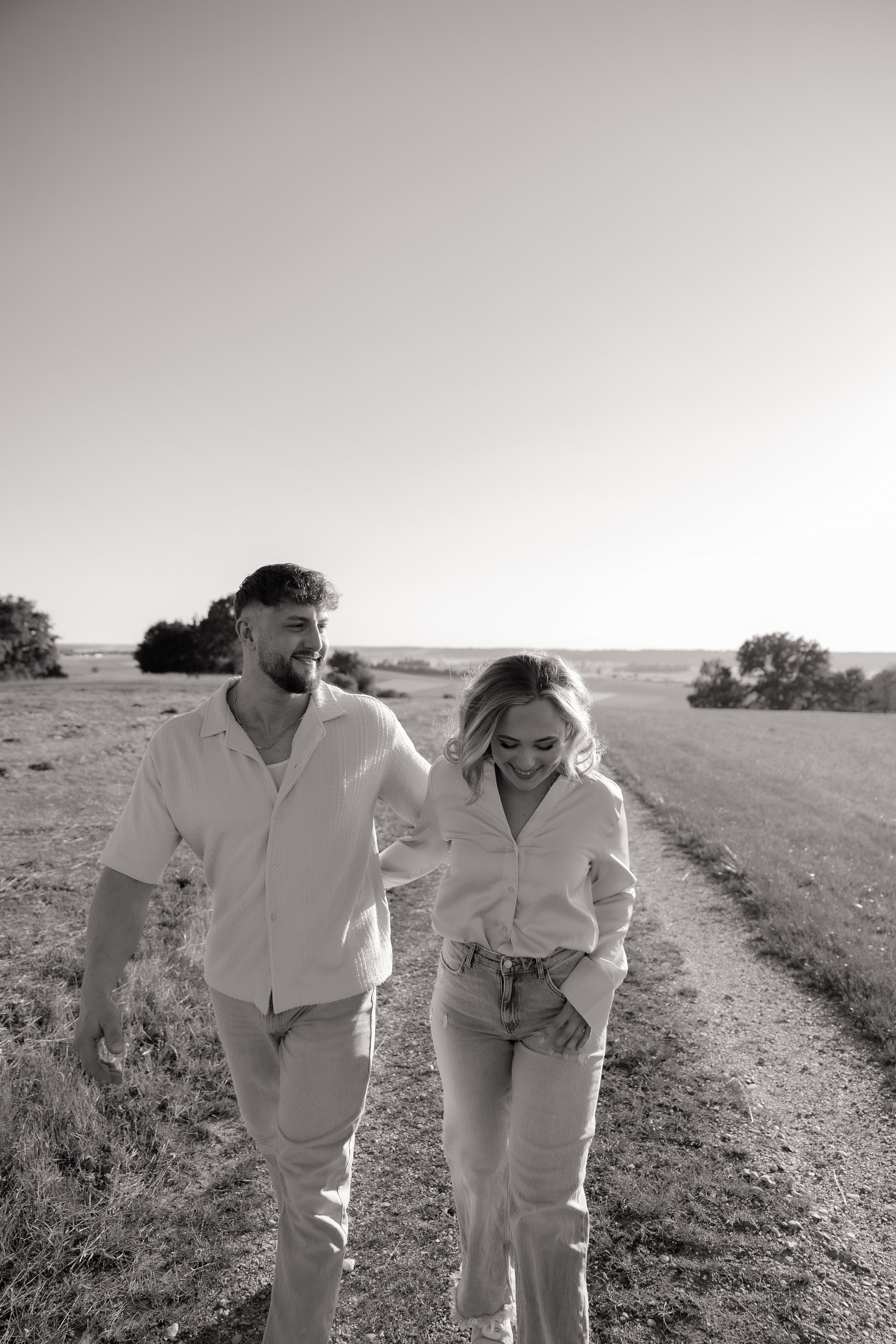 Two people laugh while walking together on a dirt path through a grassy field.
