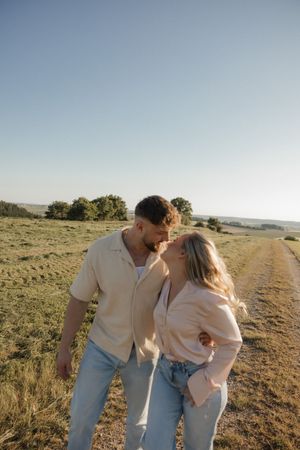 Playful couple in light colored clothing embrace on a sunlit path through rolling hills.