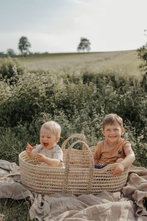 Three young children laying and playing together in a woven basket outdoors.