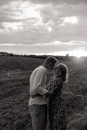Black and white photo of a couple sharing an intimate moment at sunset in a field.
