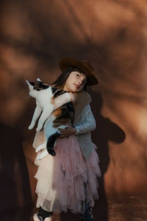 A dreamy portrait of someone in a tulle dress holding a black and white cat against a dark brown background.