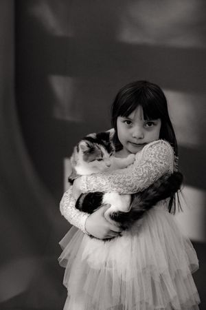 A black and white portrait showing a child in a white lace dress hugging a spotted cat.