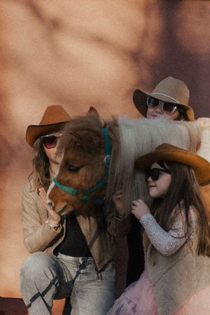 Three figures in cowboy hats pose with a pony wearing a turquoise halter against a warm brown background.
