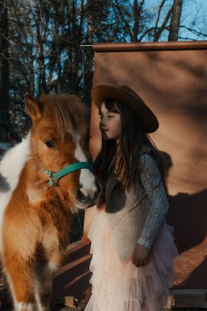 A child in a lace dress and hat stands next to a chestnut pony with a turquoise halter.