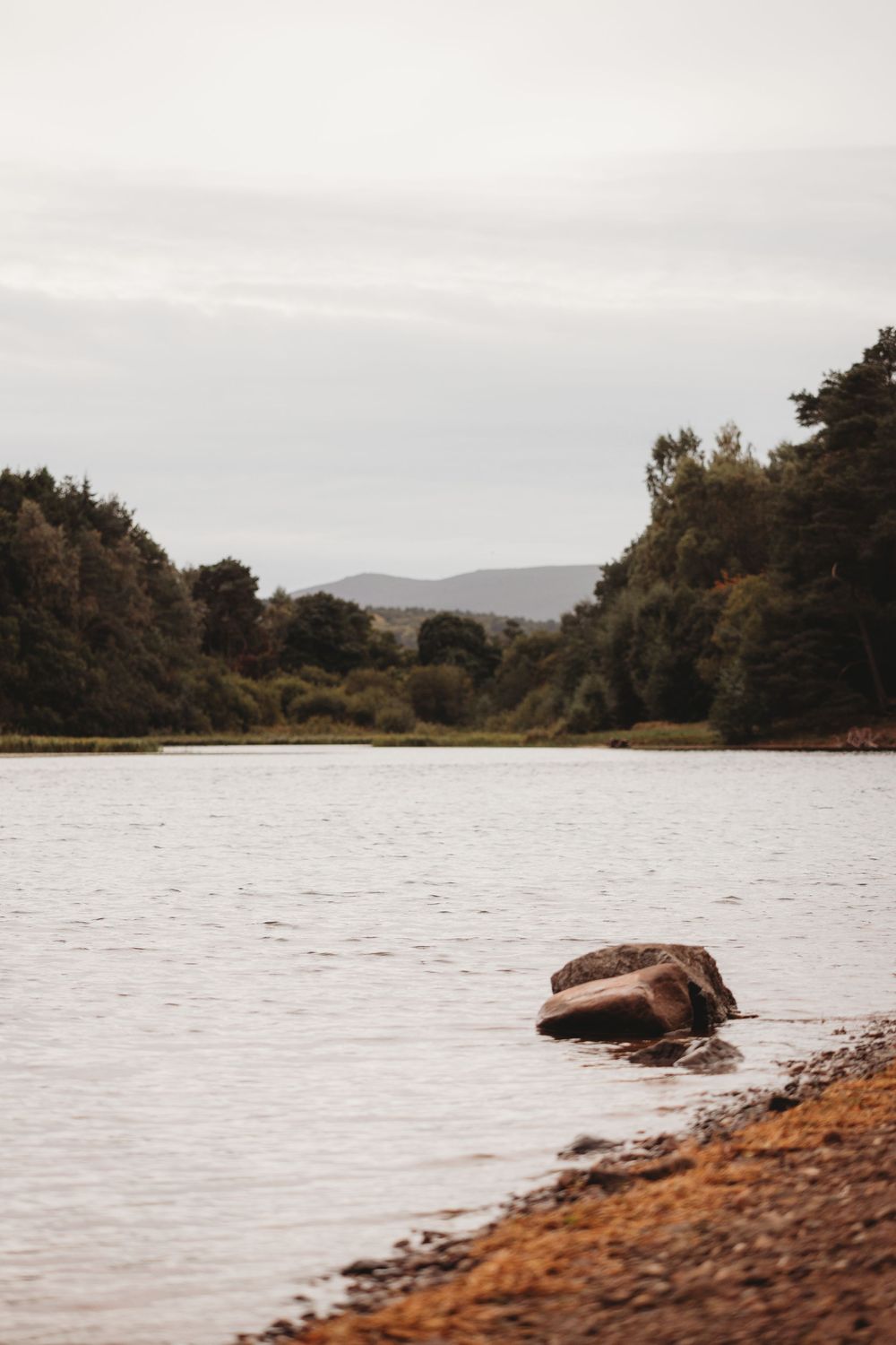 A tranquil river scene with a large rock on the pebbly shore, lush green trees, and misty hills under an overcast sky.