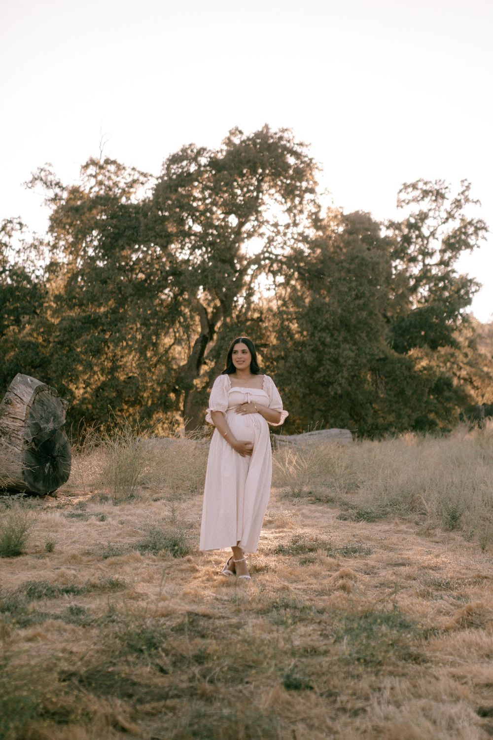 A person in a flowing white dress stands in a golden field at sunset among scattered trees and dry grass.