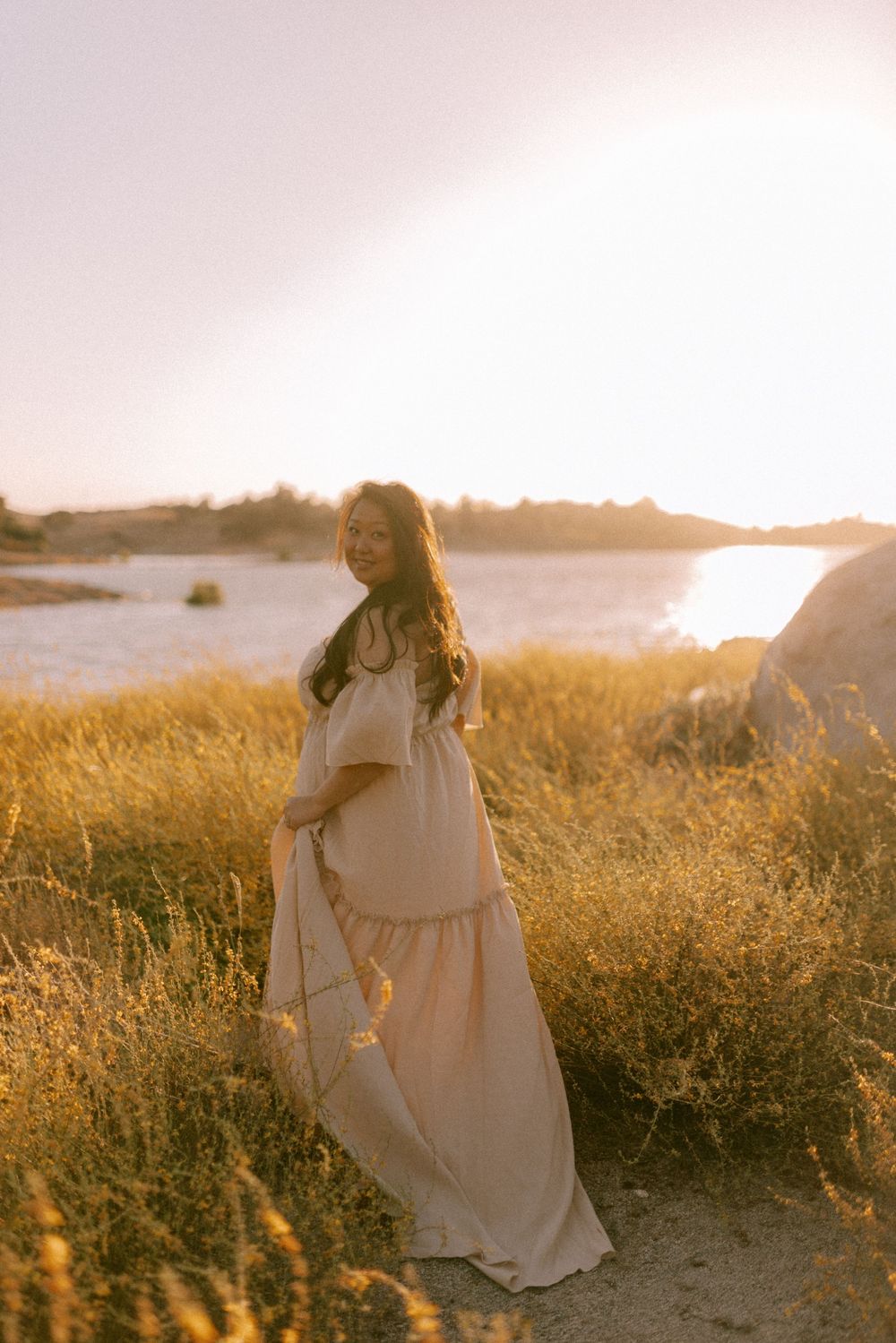 Pregnant woman in beige off-shoulder maxi dress posing in golden grass field near water at sunset.