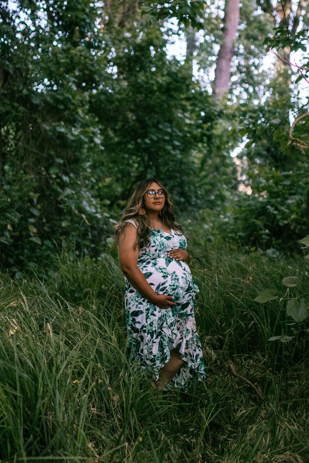 A figure in a floral dress poses gracefully among tall grass in a wooded outdoor setting during golden hour.