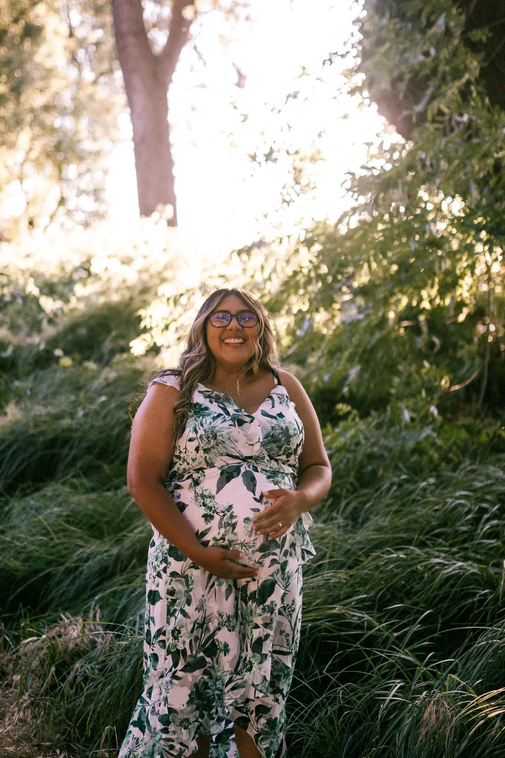 A joyful figure in a floral print dress stands laughing outdoors among tall grasses with sunlight filtering through trees.