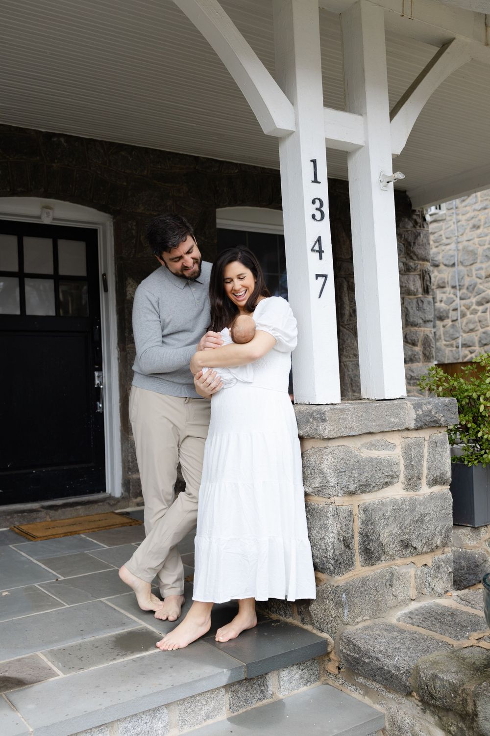 Smiling parents stand barefoot on their front porch holding their newborn baby together.