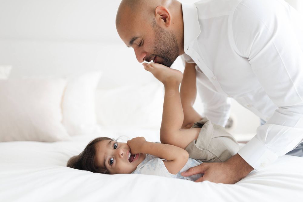 Loving father leans over his laughing baby on a white bed in a bright room.