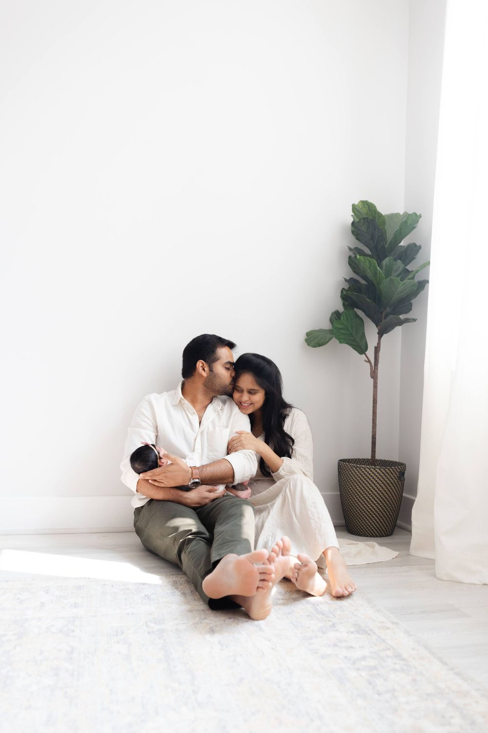 Couple sitting on floor holding newborn baby near white wall and plant.