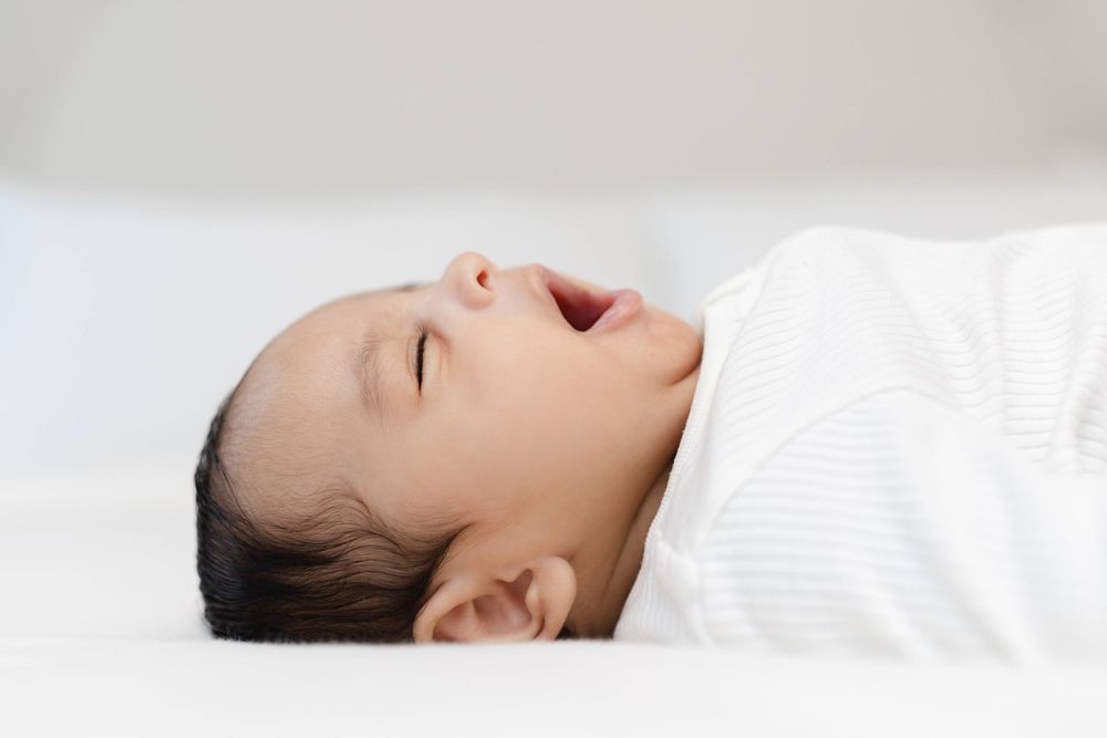 Peaceful newborn baby yawning while swaddled in white blanket.