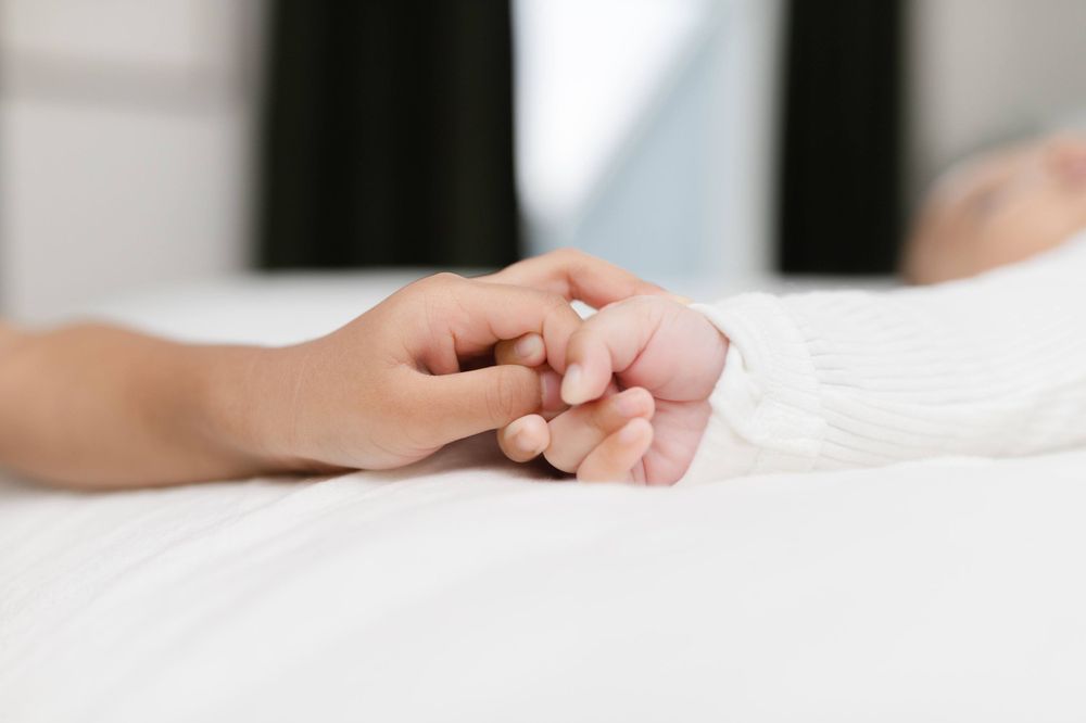 Adult hand gently holding tiny newborn baby hand on white surface.