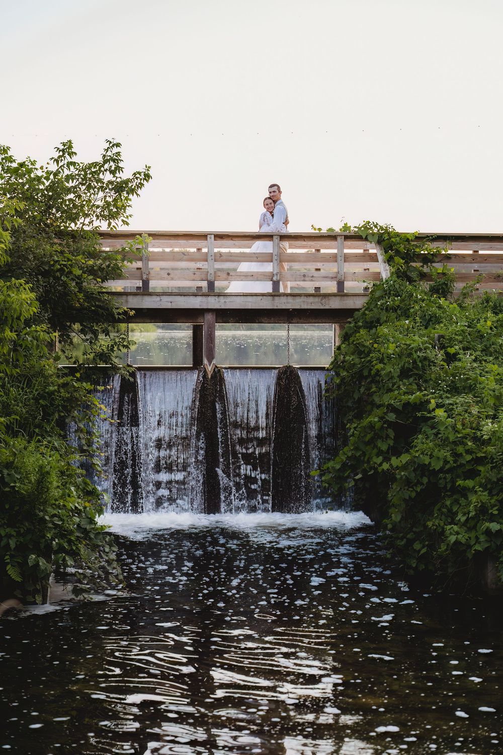 A small waterfall cascades over a concrete dam structure surrounded by green foliage and reflecting pond water.
