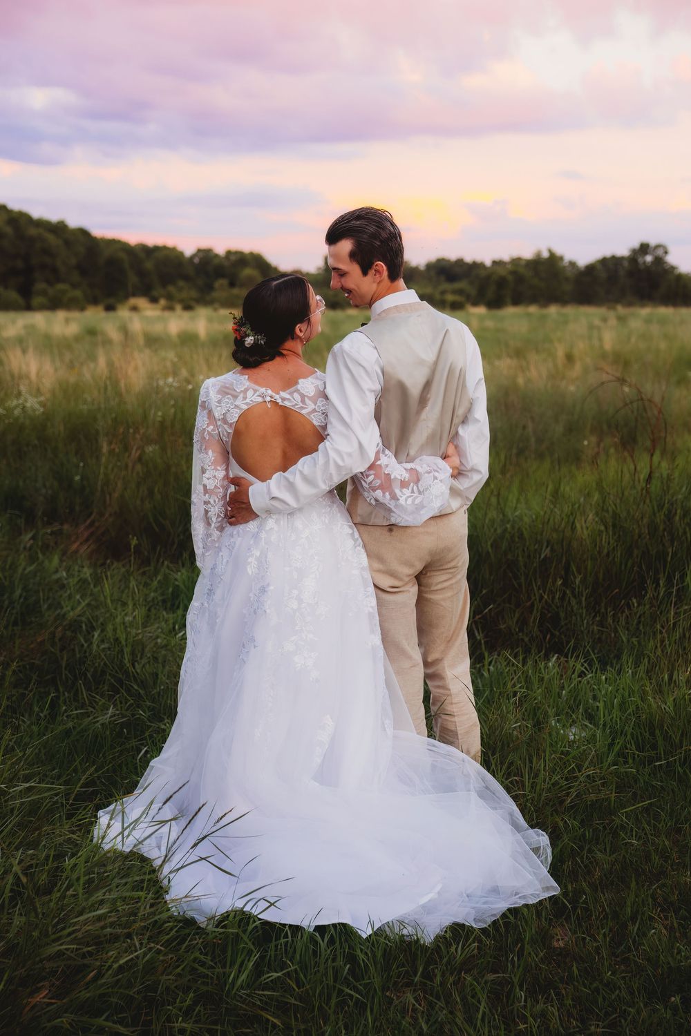 A couple in wedding attire embraces while standing in a grassy field at sunset.