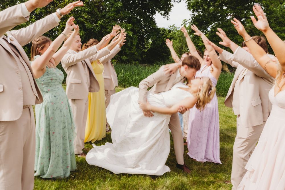 Bride and groom share a romantic dip kiss surrounded by cheering wedding party on a lush green lawn.