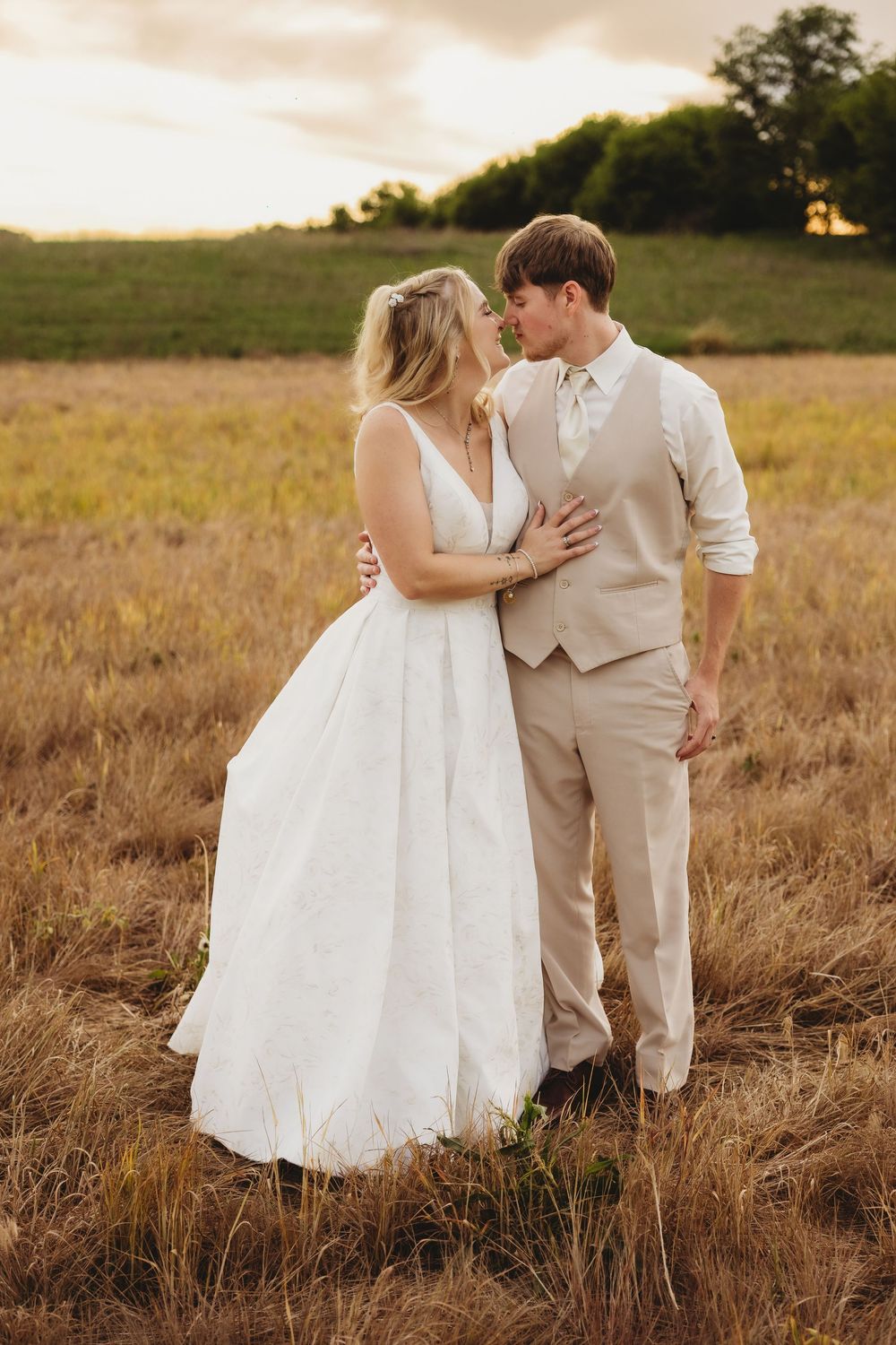 Bride and groom sharing a romantic moment in a golden field at sunset, bride in white gown, groom in tan suit.