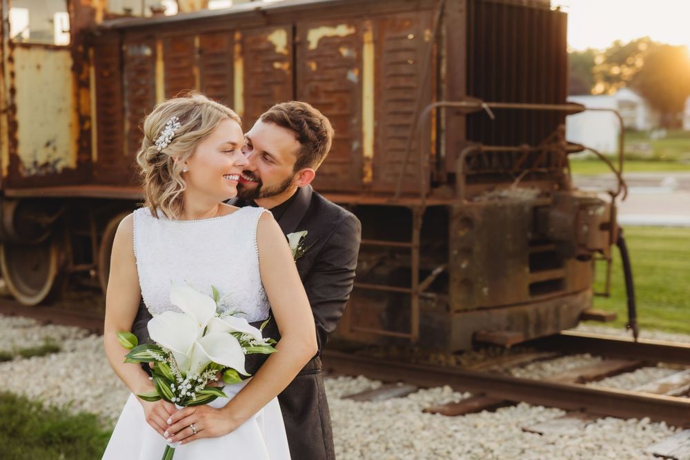 Bride and groom share a romantic moment in front of a rustic train car, bride holding white bouquet.