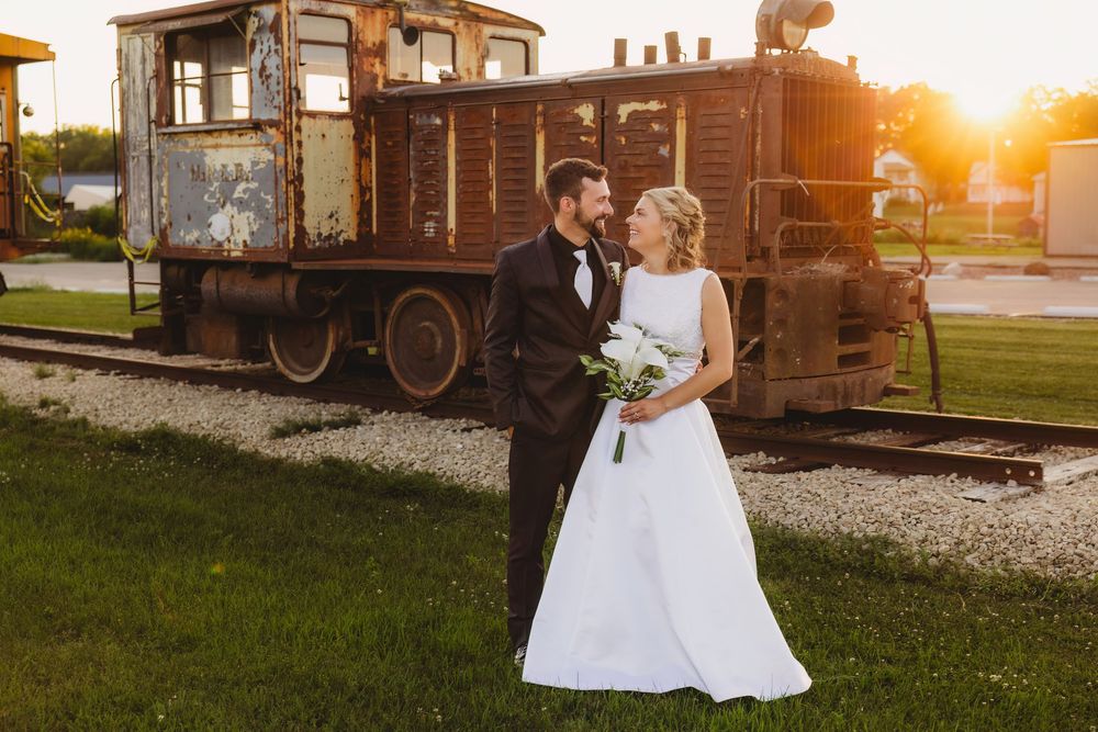 Bride and groom pose before a rustic vintage train at golden hour, bride holding white bouquet in elegant gown.
