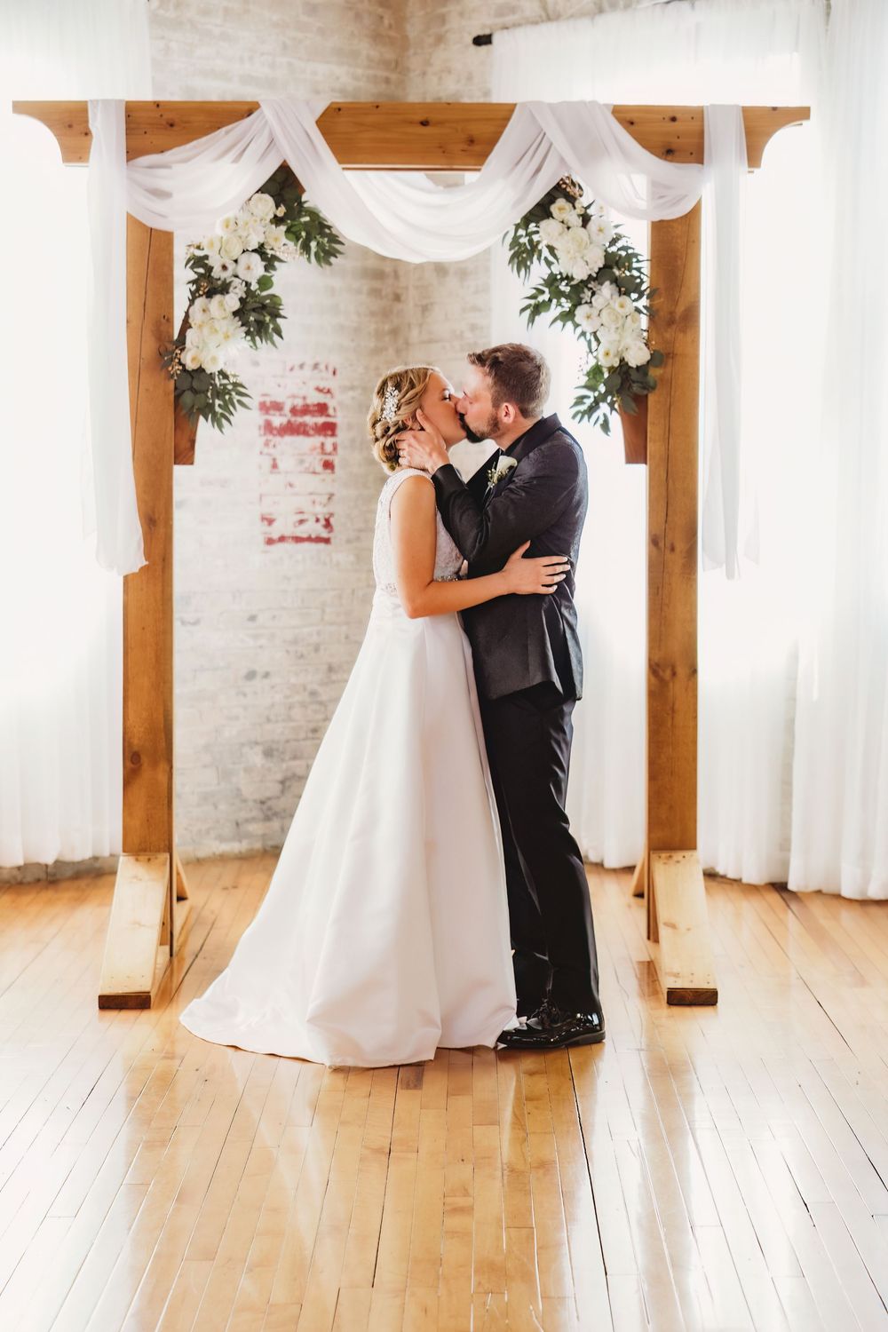 Bride and groom share their first kiss under a rustic wooden arch adorned with white draping and floral arrangements.