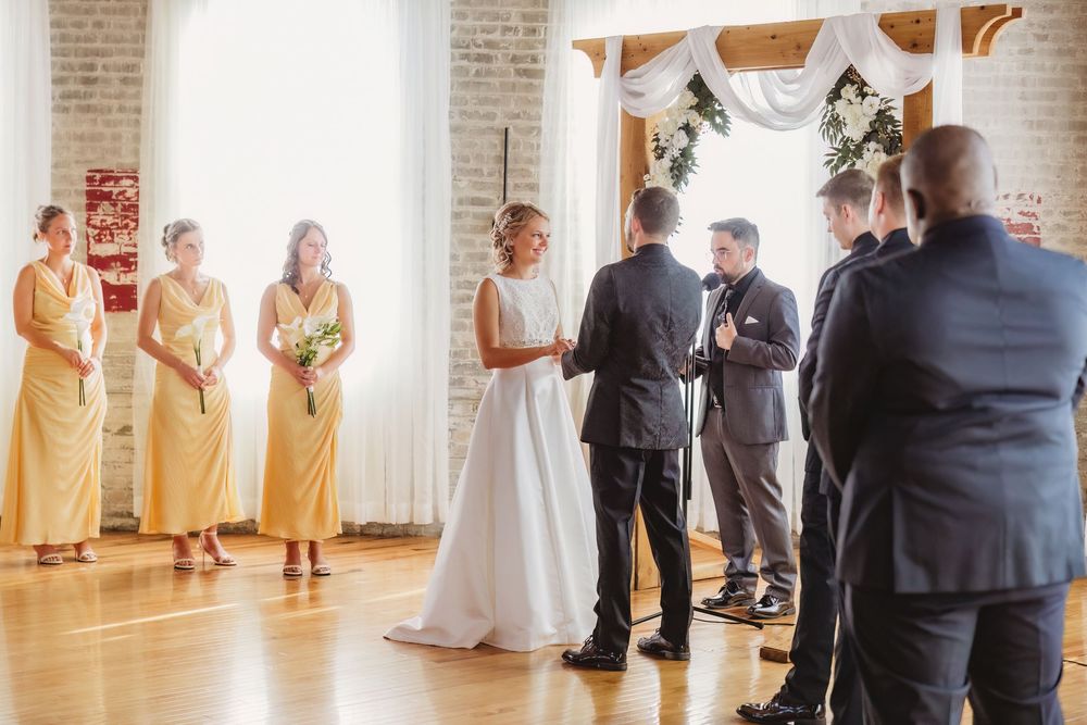 Bride and groom exchange vows at indoor wedding ceremony with bridesmaids in yellow dresses and groomsmen by floral arch.
