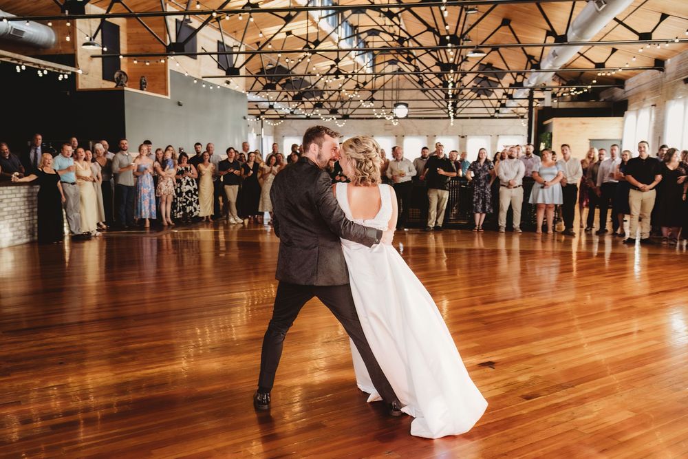 Bride and groom share first dance on wooden floor while wedding guests watch in elegant industrial venue.