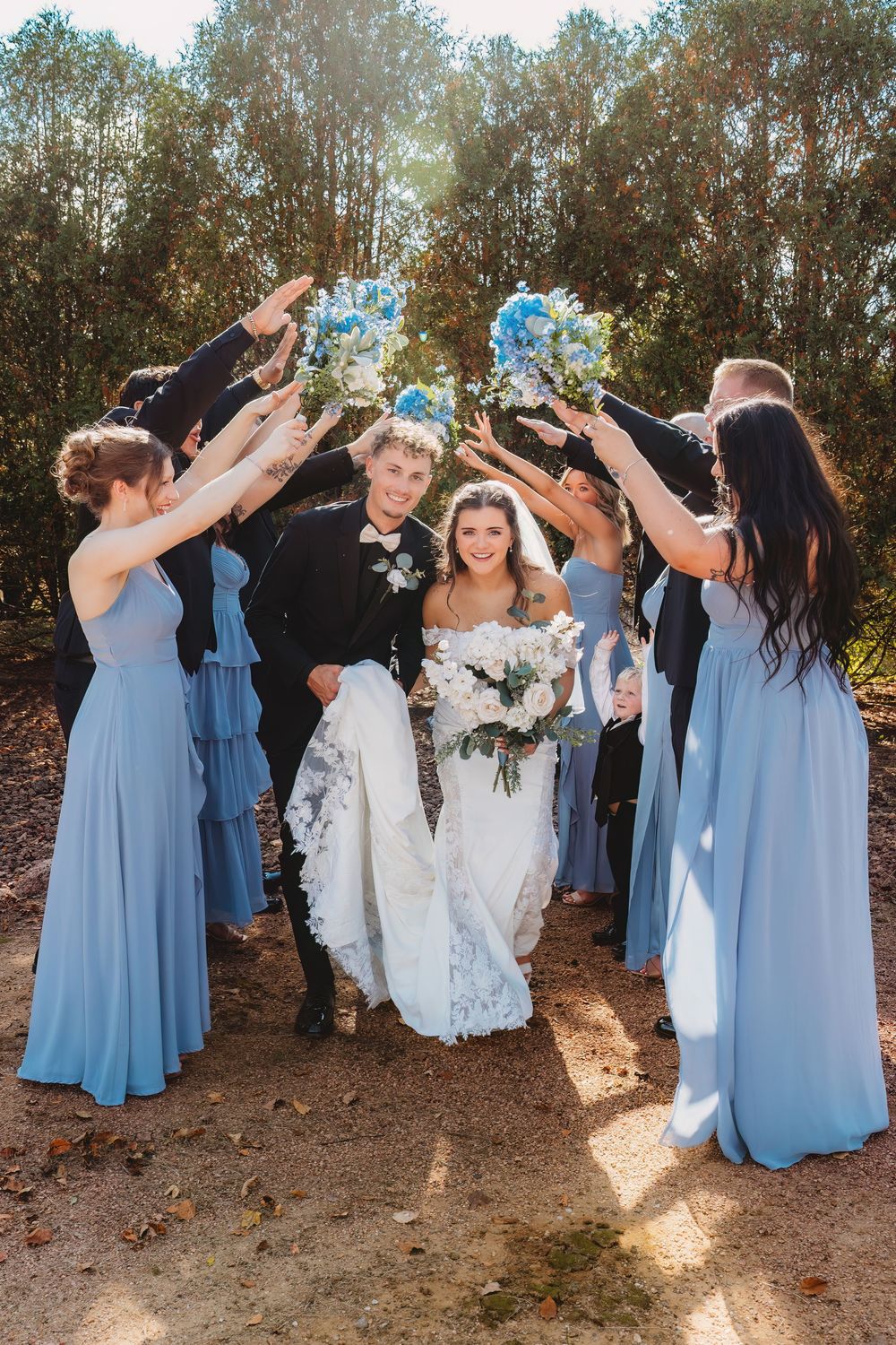 Wedding party celebrates outdoors with bridesmaids in light blue dresses forming an arch on a sunny forest path.