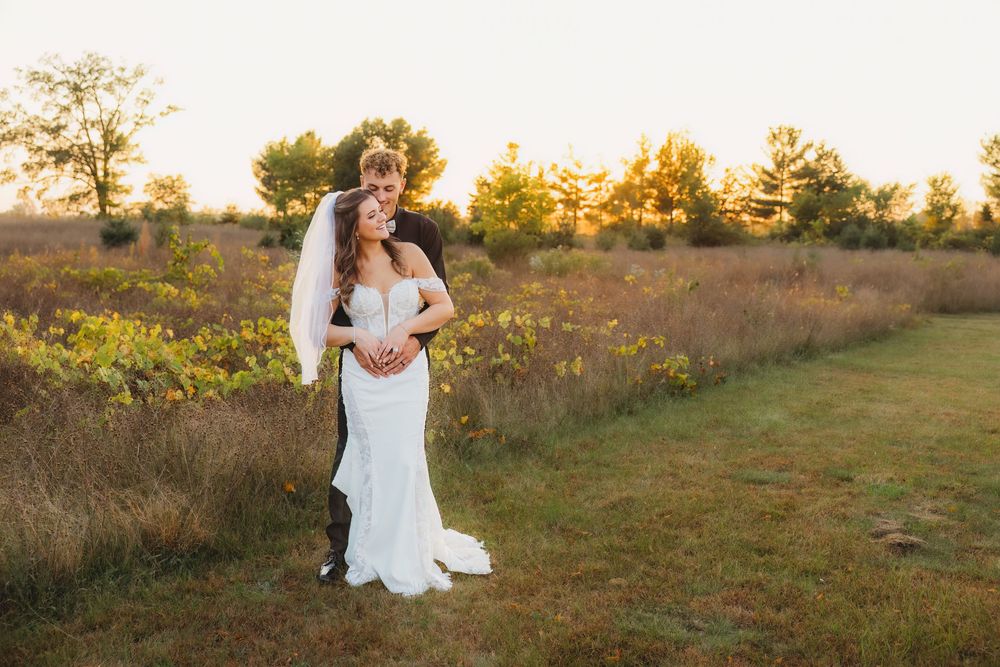 A couple embraces in a golden sunlit field during sunset while wearing wedding attire.