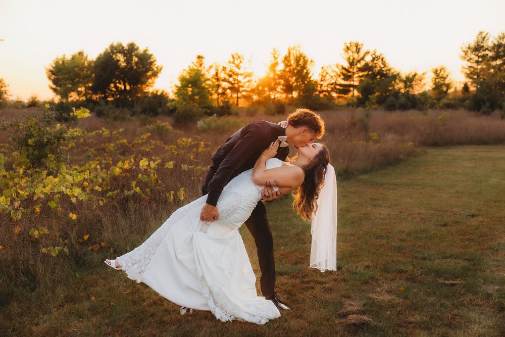 A couple in wedding attire embraces in a golden field during sunset while trees line the horizon.