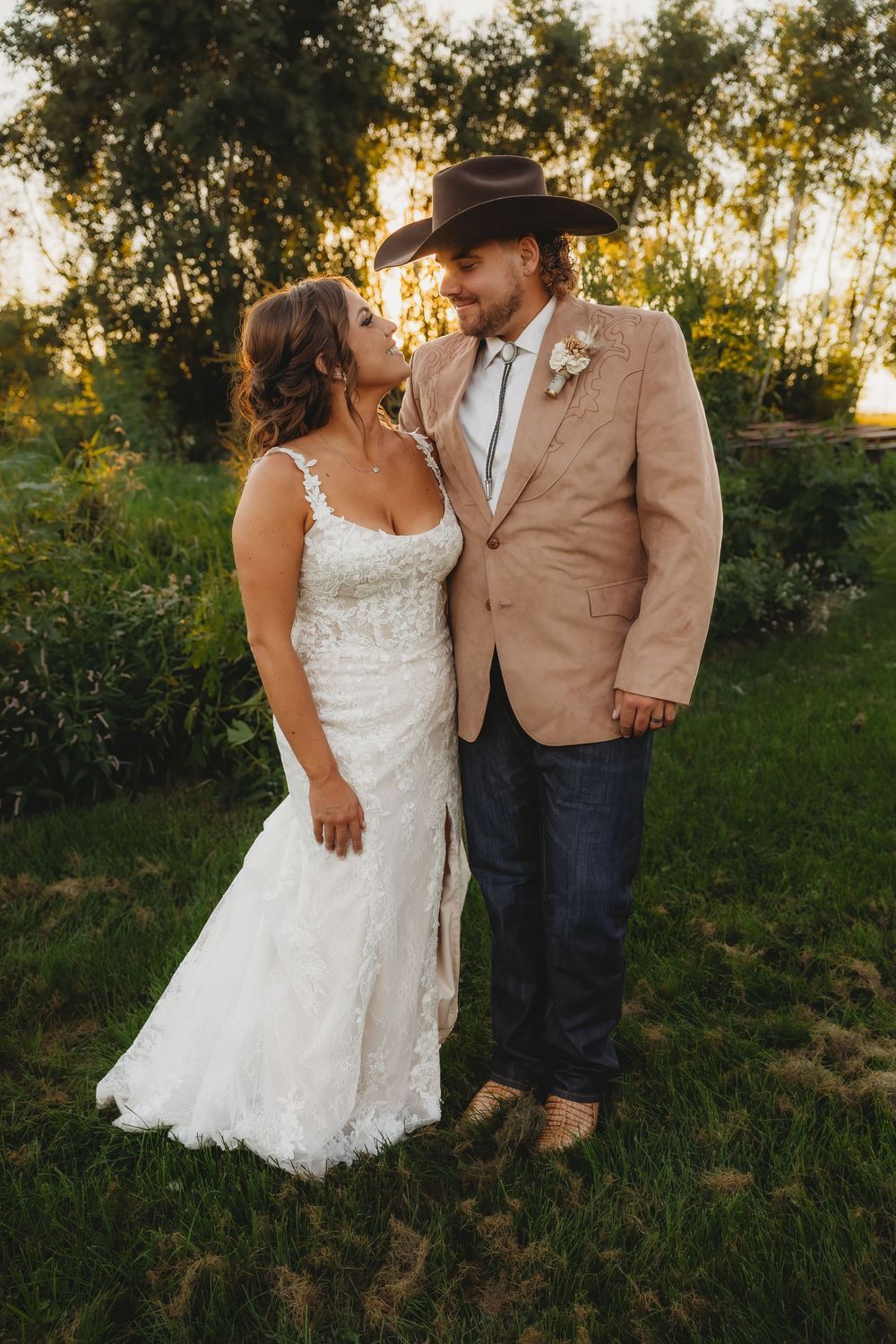 A romantic sunset wedding photo of a couple in a white lace gown and tan suit with cowboy hat standing in a grassy field.
