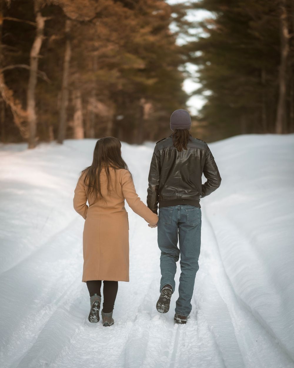 Couple holding hands walking down a snowy forest road in winter, woman in tan coat and man in leather jacket.