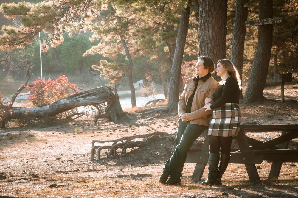 A couple stands together in a romantic autumn forest setting with sunlight filtering through the trees.