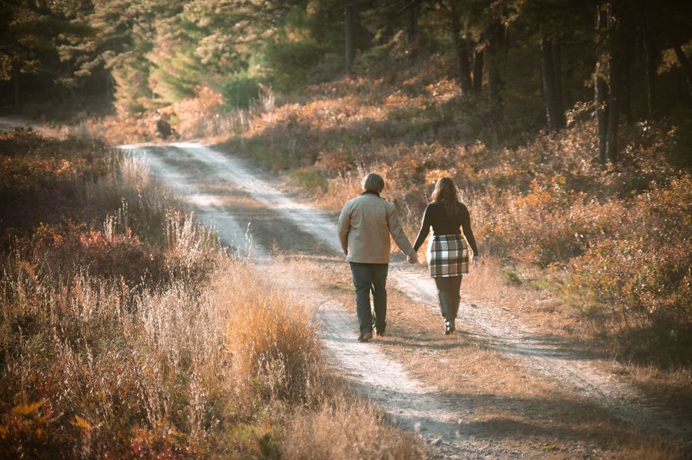 Two people walk down a winding autumn path through golden grass and trees at sunset.