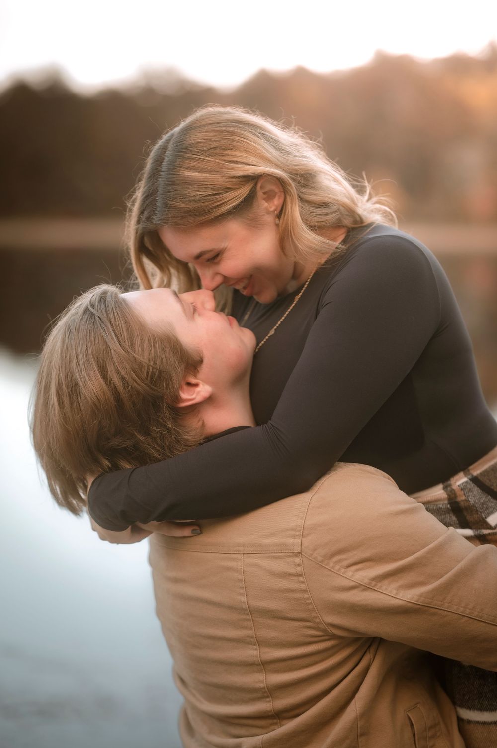 A romantic couple embraces and kisses during sunset by a lake with warm golden lighting.