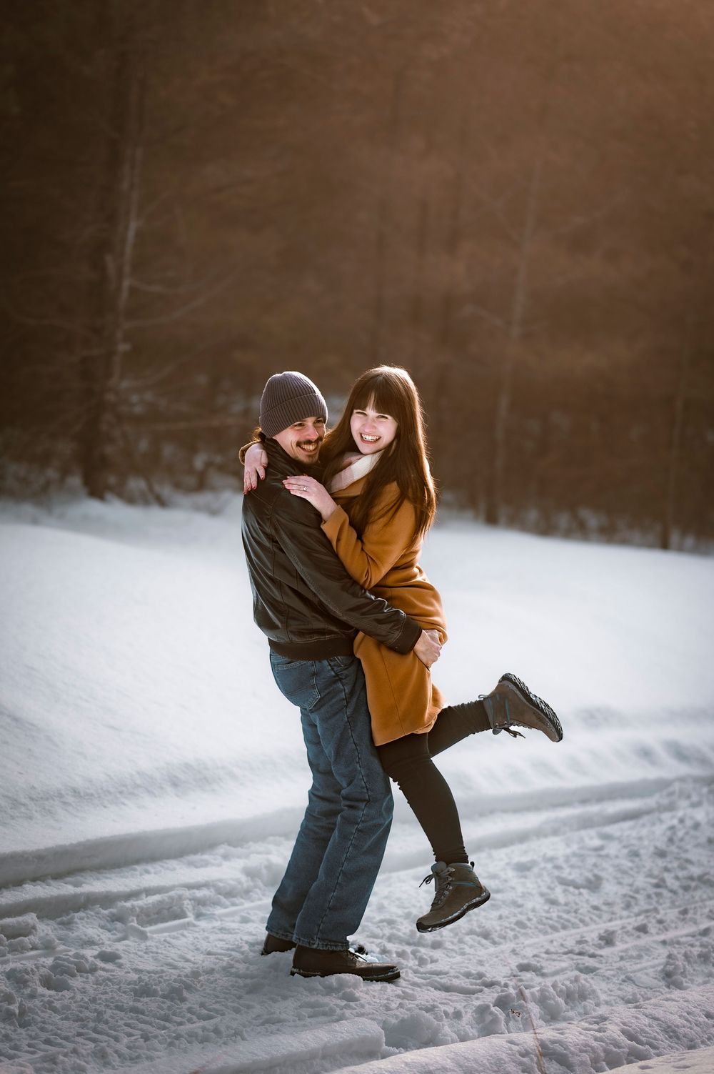 Couple embracing and laughing in snowy winter landscape, woman in brown sweater lifted by man in gray beanie.