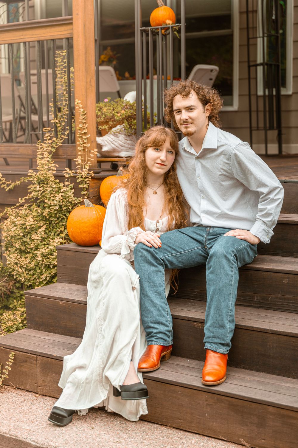 A couple sits on wooden steps decorated with autumn pumpkins outside a rustic building.