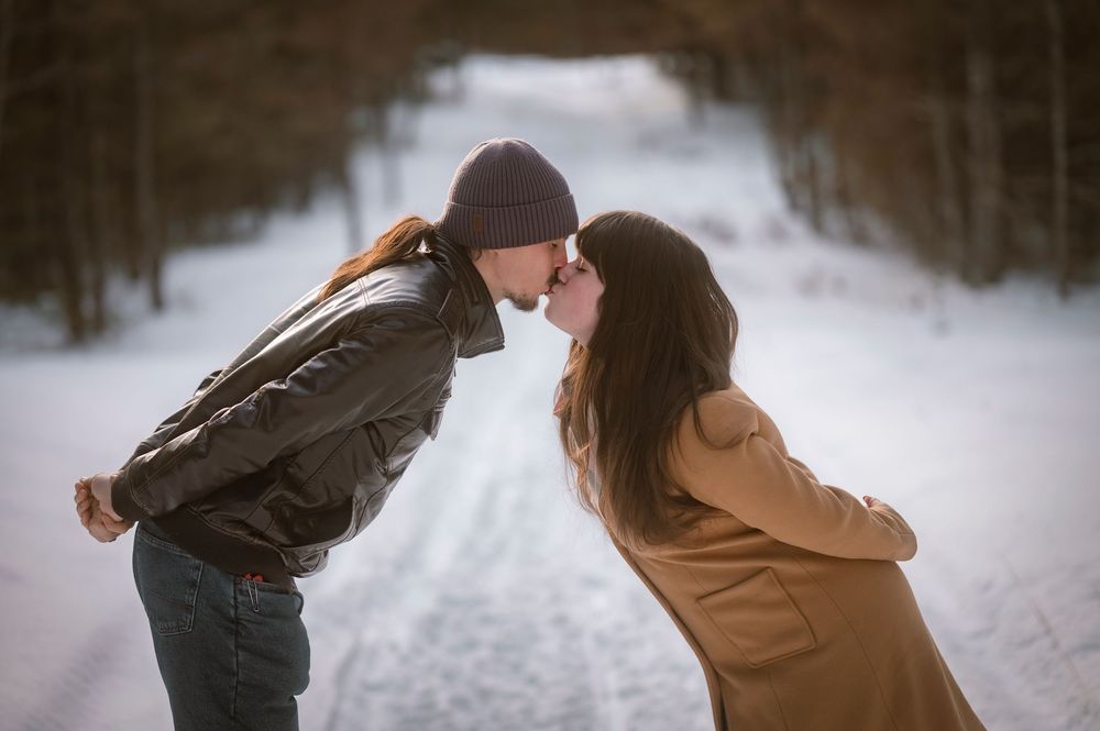 Couple sharing a romantic kiss on a snowy winter road surrounded by bare trees.