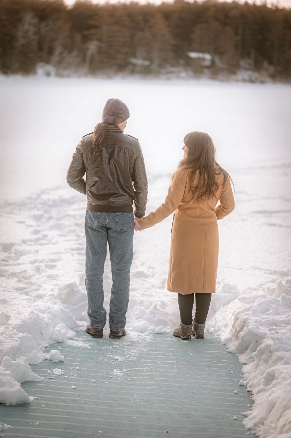 Couple holding hands on a snowy dock, facing a frozen lake in winter, wearing warm coats and boots.