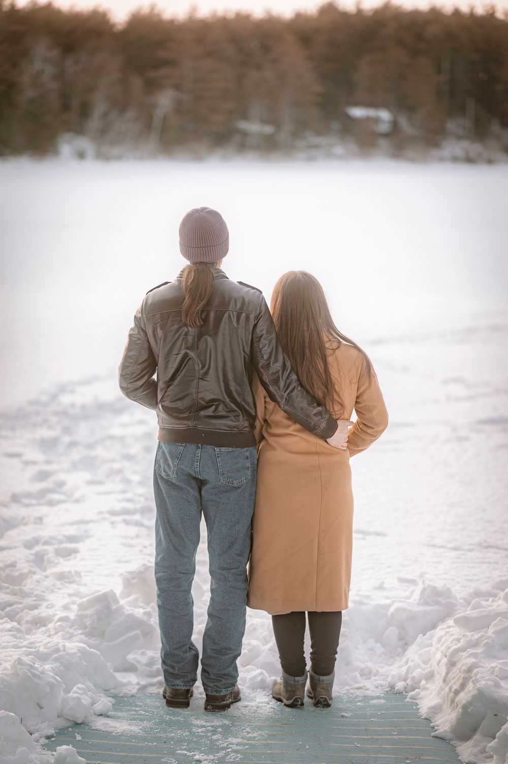Couple standing together on a snowy dock, embracing while gazing at a frozen lake surrounded by winter trees.