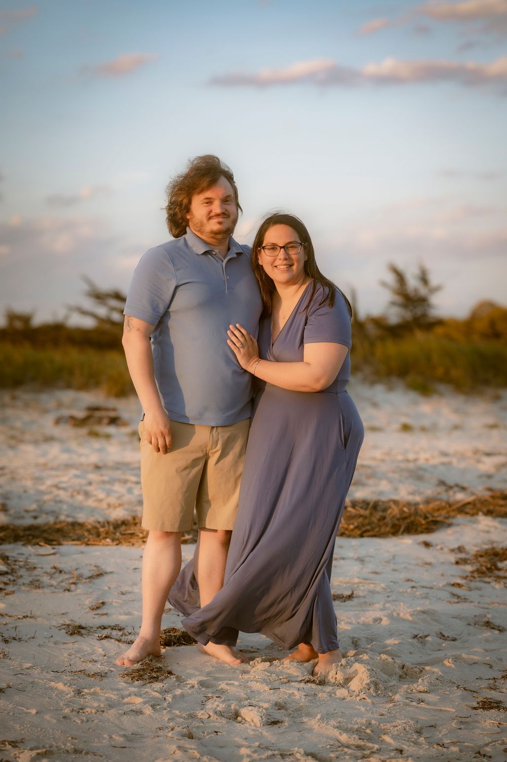 Couple posing together on sandy beach at golden hour, woman in blue maxi dress, man in blue polo and khaki shorts.
