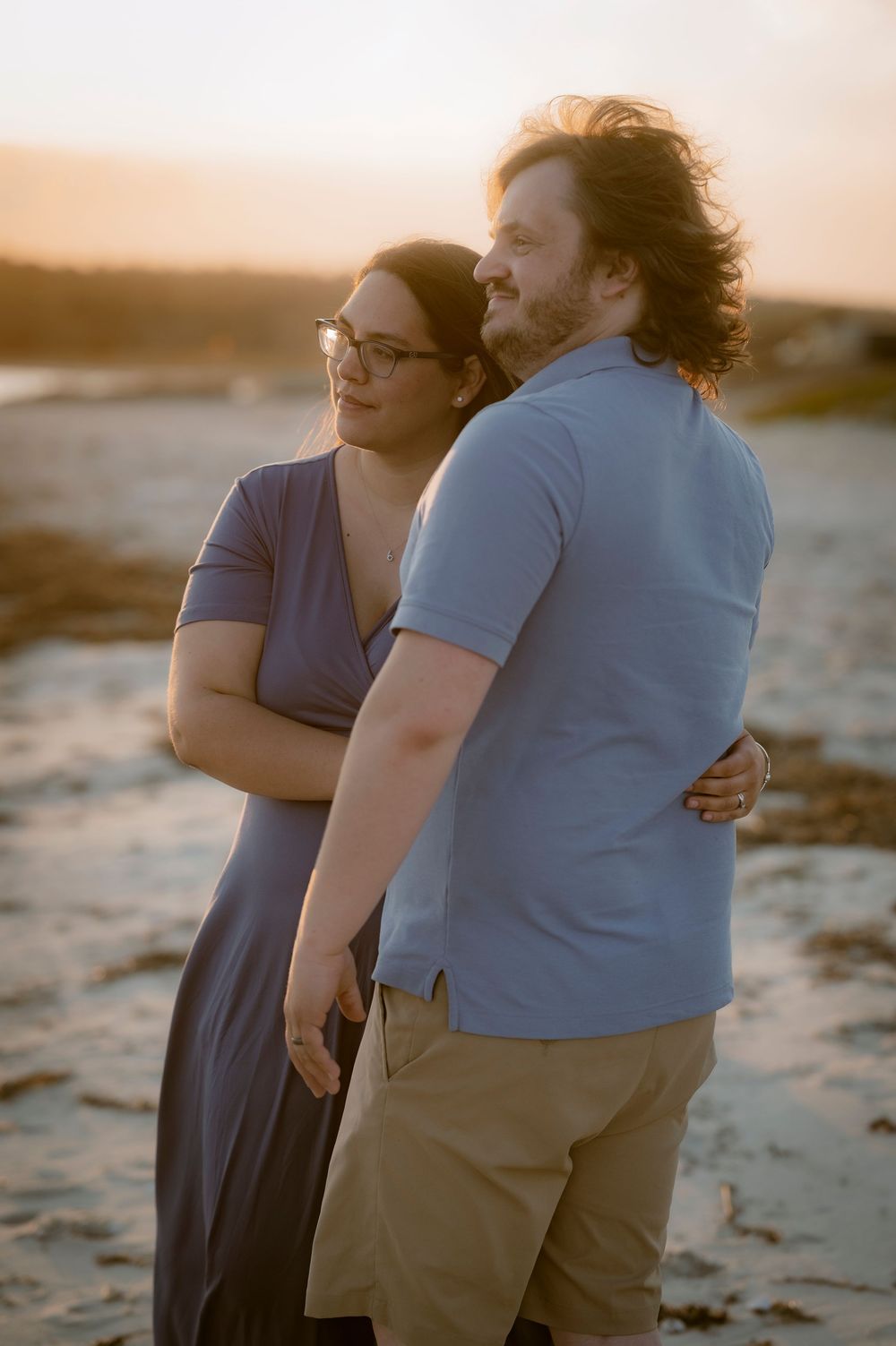Couple embracing at sunset on a beach, woman in blue dress with glasses, man in light blue polo shirt.
