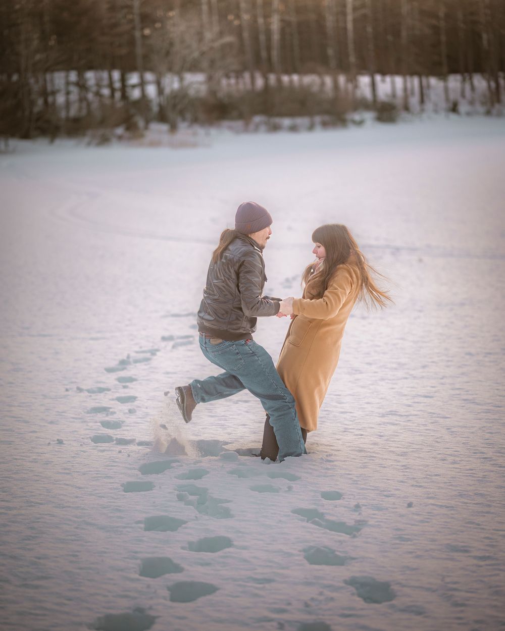 Couple dancing playfully in a snowy winter landscape, leaving footprints in the snow with trees in the background.