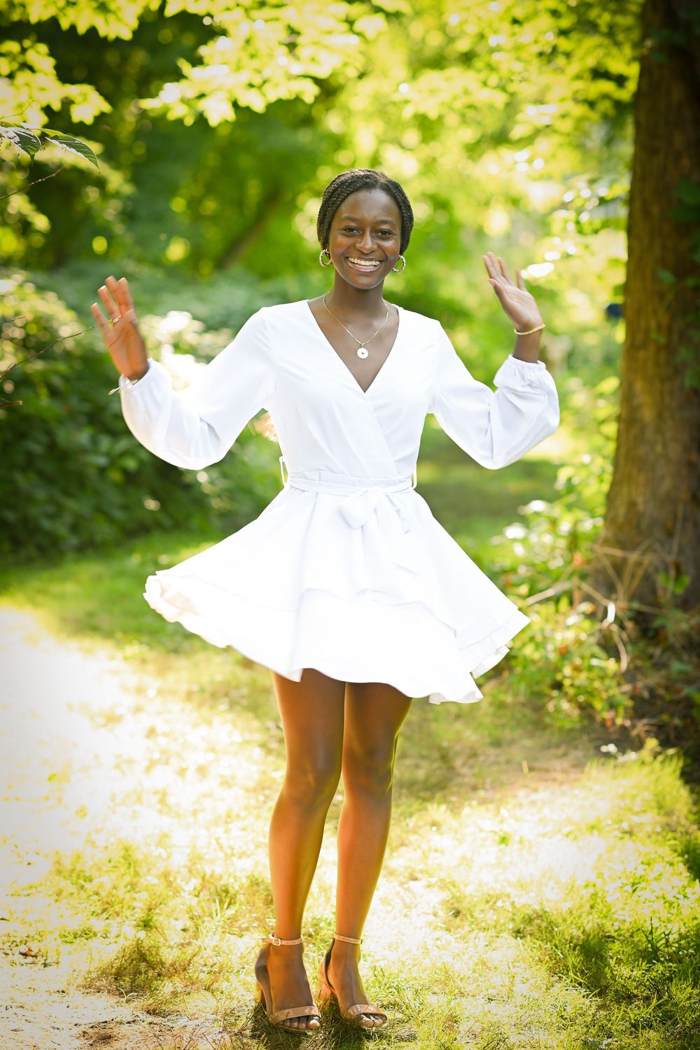 Young Black woman in white dress dancing on a sunlit garden path surrounded by lush green trees.
