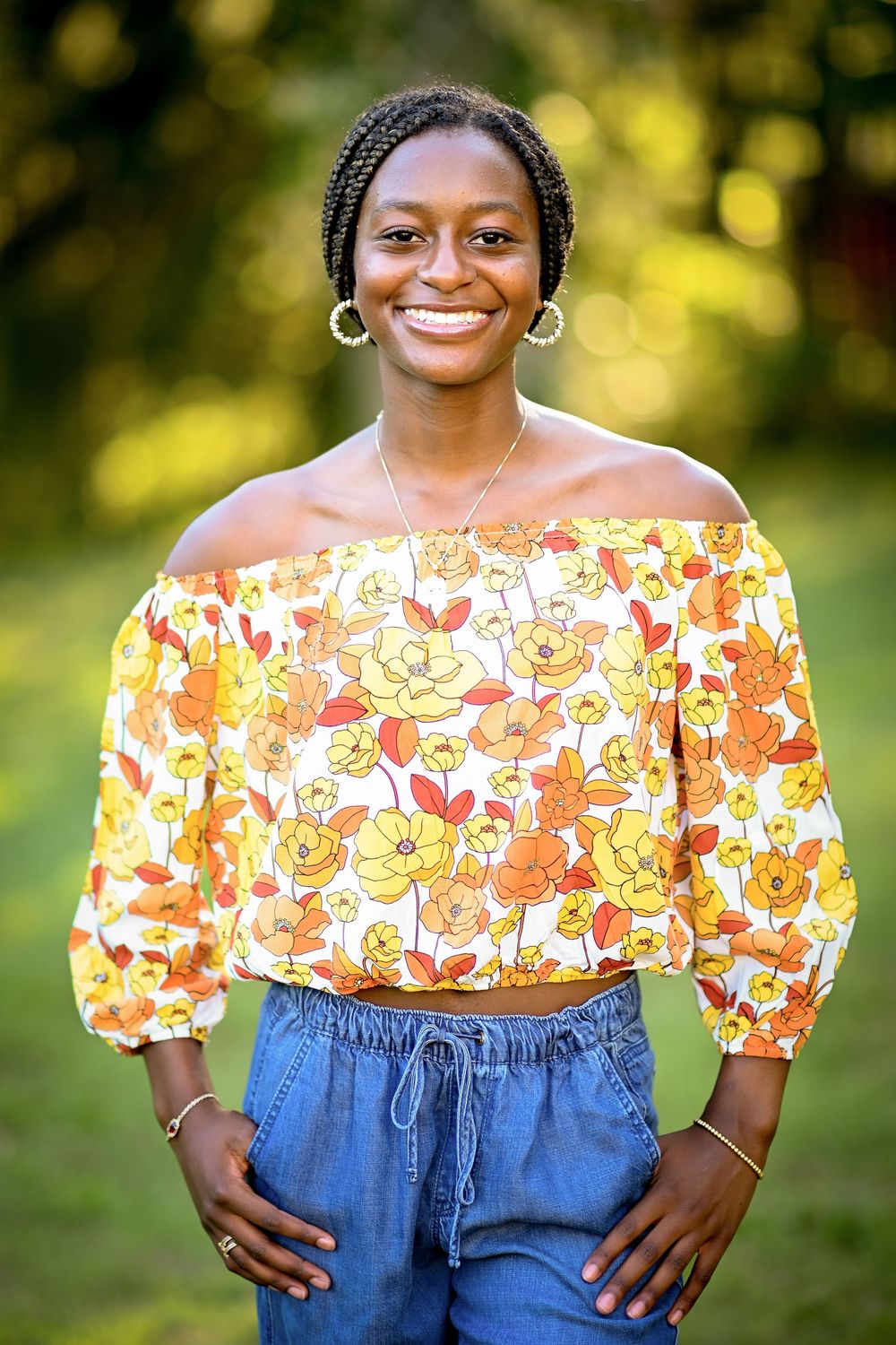 Smiling young woman in floral off-shoulder top and jeans posing outdoors in a lush green park setting.