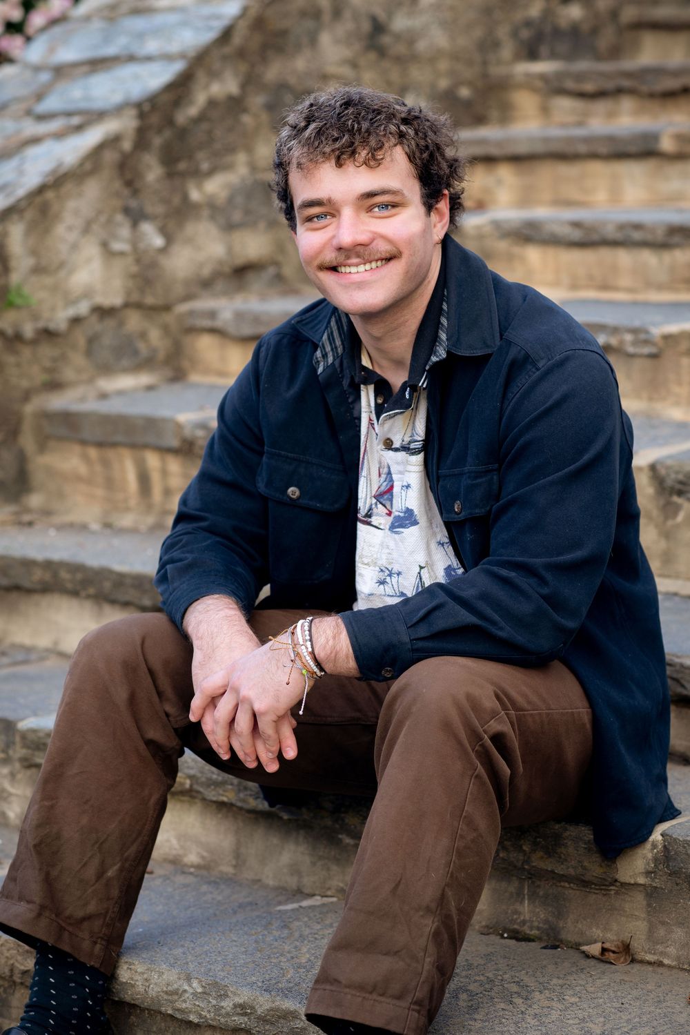 Smiling young man with mustache sitting on stone steps outdoors, wearing navy jacket and brown pants.